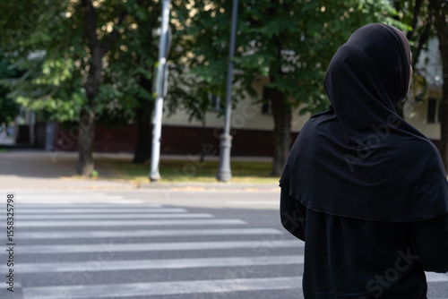 Woman in black attire stands at crosswalk observing surroundings in a park area on a sunny day