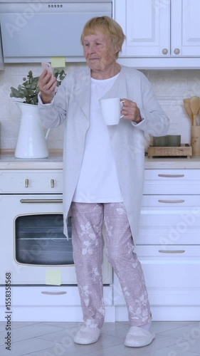 Vertical screen elderly woman enjoying morning tea while browsing smartphone in bright, modern kitchen, showcasing active senior lifestyle and digital connectivity