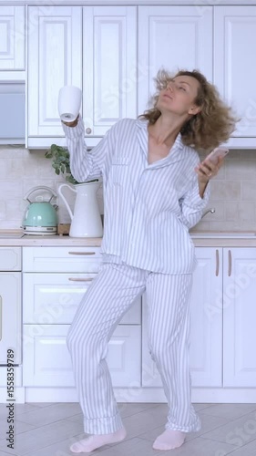 Vertical screen happy woman wearing pajamas dances and sings while holding a smartphone and a coffee cup in a bright kitchen, enjoying her morning routine