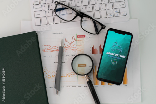 A smartphone displaying Agriculture Market Outlook report is placed atop a stack of papers with graphs and charts, accompanied by yellow notebook, keyboard, glasses and magnifying glass 