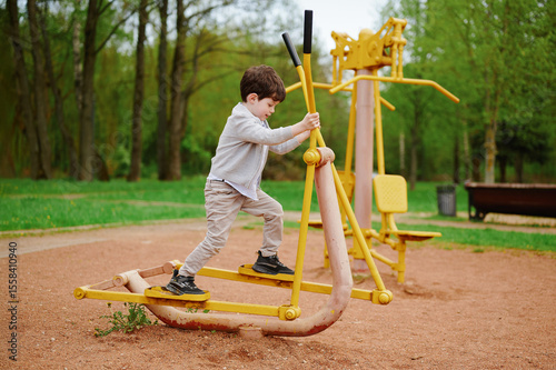 child playing outdoors on exercise equipment in park setting. young boy balancing on fitness apparatus surrounded by trees. exploring outdoor activities and healthy lifestyle concepts