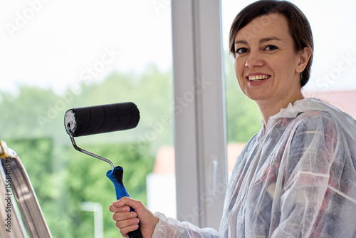 Smiling woman in protective overalls holds paint roller while painting walls in room. Home renovation and interior upgrade concept