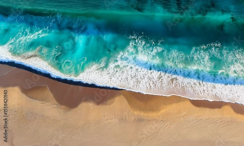 Aerial view shows turquoise ocean waves crashing onto a sun-drenched sandy beach. Shadows and textures create depth