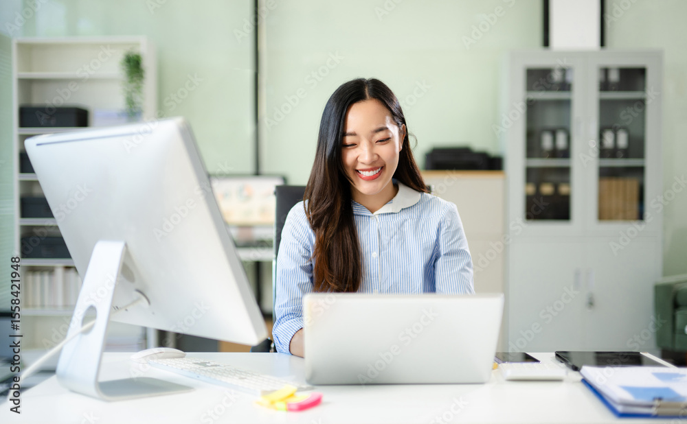 Happy Asian businesswoman reviews financial reports at her office desk. Bright workspace, confidence, success Happy Asian businesswoman reviews financial reports at her office desk. Bright workspace, confidence, success