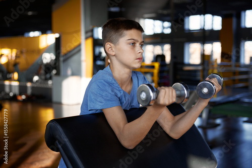 Teenage boy pumping biceps enjoying training with dumbbells at gym