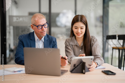 Senior businessman and young businesswoman are working together in a modern office, using a laptop and a tablet to analyze data and discuss business strategies