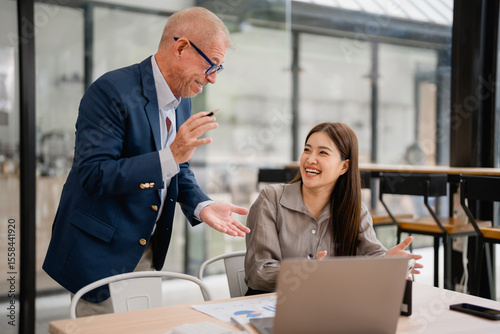 Senior manager coaching a young asian businesswoman using laptop in a modern office, they're smiling and discussing about work, she's wearing a gray shirt and he's wearing a blue suit