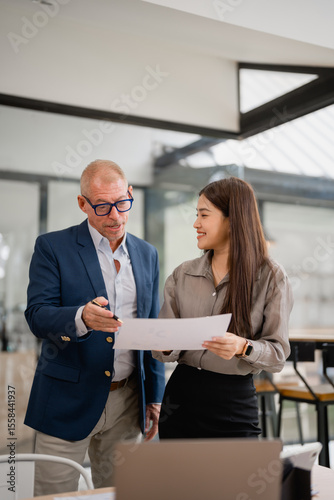 Senior manager explaining marketing strategy to a young businesswoman holding documents, discussing a project in a modern office, collaborating and working together to achieve business goals