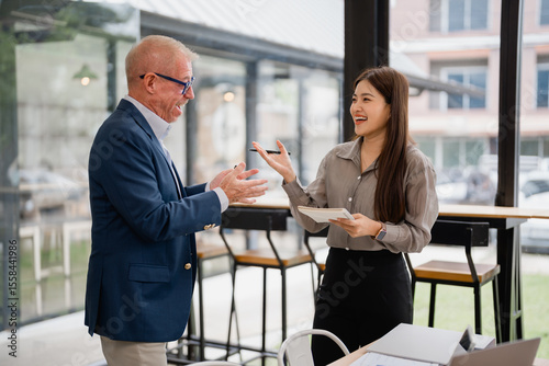 Senior manager and young businesswoman standing and discussing work together with happy emotion, they are laughing during a meeting in a modern office