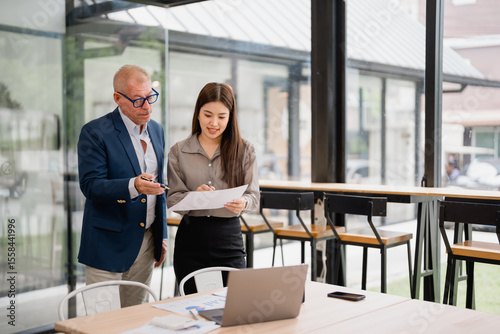 Senior manager explaining marketing strategy to young businesswoman holding documents and pointing at laptop in modern office with big windows