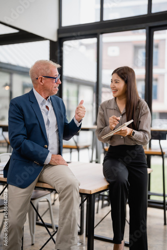 Senior manager showing thumbs up during a discussion with a young businesswoman taking notes, sitting on a table in a modern office, collaborating and sharing ideas for business success