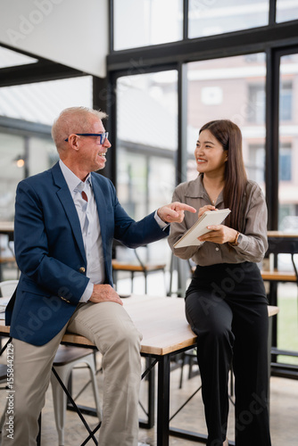 Senior manager pointing at a notebook while discussing strategies with a young Asian businesswoman, who is taking notes in a bright, modern office filled with natural light