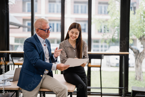 Senior manager explaining marketing strategy to young asian businesswoman while analyzing statistics report, pointing at charts and discussing project details in modern office
