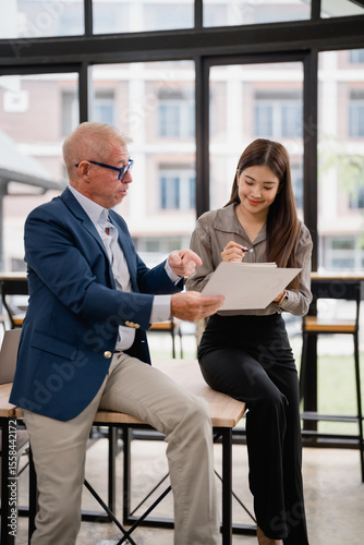 Senior manager explaining project details to young asian businesswoman taking notes while sitting on a table in modern office during a working meeting