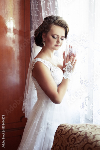 A woman in a white dress is standing in front of a window. She is wearing a veil and has her hands on her hips. Scene is elegant and romantic