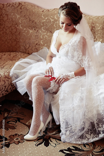 A bride is sitting on a couch in her wedding dress. She is wearing white stockings and a red ribbon