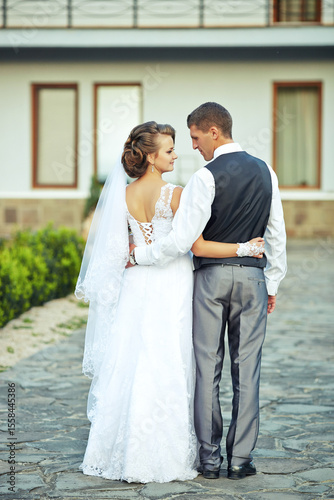 A bride and groom are standing on a stone walkway, posing for a picture. The bride is wearing a white dress and the groom is wearing a black vest. Scene is happy and celebratory
