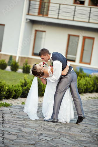 A bride and groom are kissing on a cobblestone walkway. The bride is wearing a white dress and the groom is wearing a gray suit