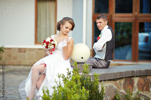 A bride and groom are sitting on a stone wall, with the bride holding a bouquet of flowers. The man is wearing a vest and the woman is wearing a white dress. Scene is romantic and intimate