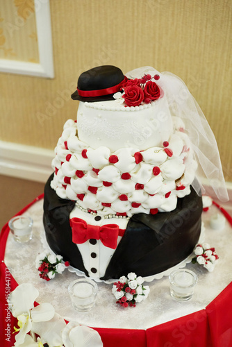 A wedding cake with a black and white suit and a red bow tie. The cake is decorated with red and white frosting and has a hat on top
