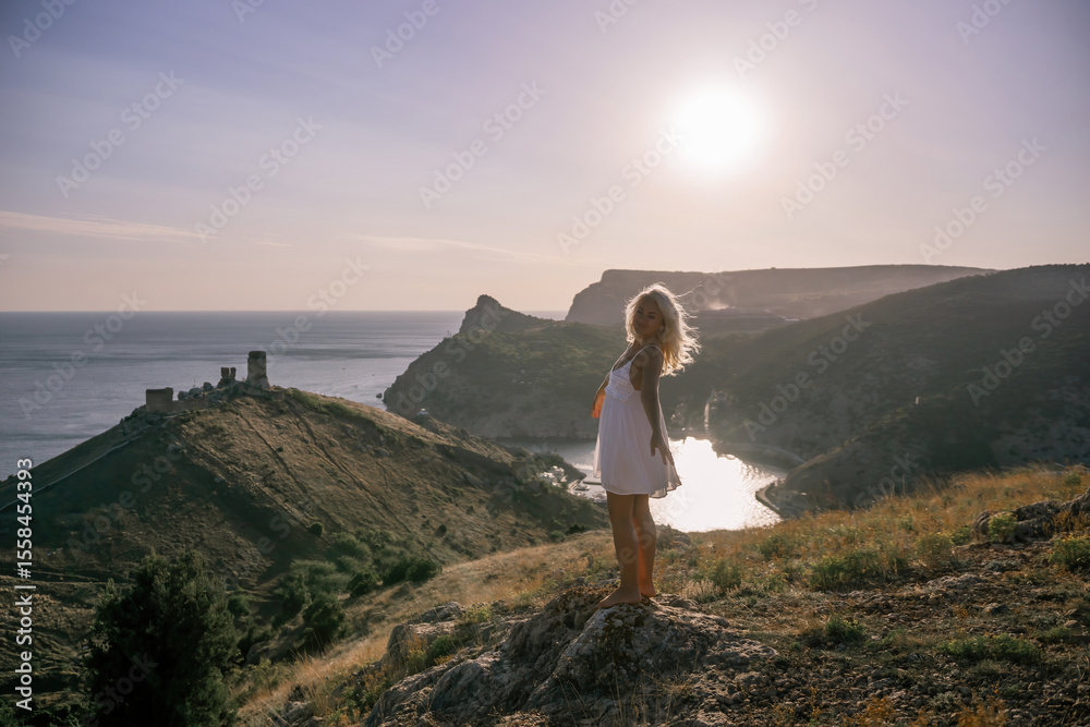 Fototapeta premium A woman stands on a hill overlooking a body of water. The sky is clear and the sun is shining brightly. The woman is enjoying the view and the peaceful atmosphere.