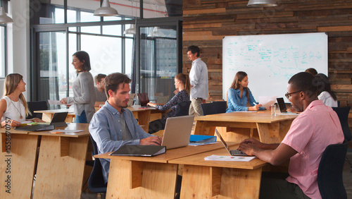 Business Team Sitting At Desks In Open Plan Office Working At Laptops