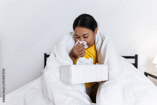 Sick woman sitting on bed wrapped in a blanket blowing nose in a tissue