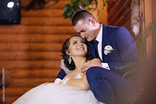 A bride and groom are sitting together on a bed. The bride is wearing a white dress and the groom is wearing a blue suit. They are both smiling and seem to be enjoying each other's company