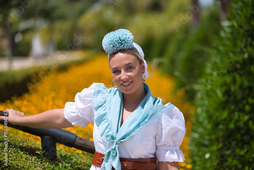 Portrait of young, beautiful, blonde woman, with typical Andalusian flamenco costume to go on pilgrimage in white color with light blue shawl and flower, leaning on a railing in an outdoor park.