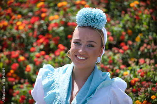 Portrait of young woman, beautiful, blonde, with typical Andalusian flamenco costume to go on pilgrimage in white color with light blue shawl and flower, with a background of red and yellow flowers.