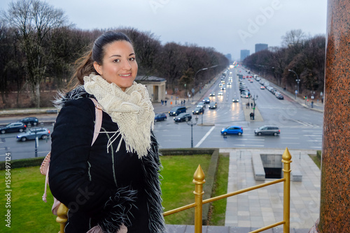Photography Young woman in winter outfit smiling at the camera, standing at the Victory Column in Berlin Germany with a busy street, cars and leafless trees in the background