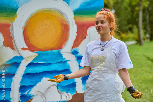 Young female with red hair discusses vibrant ocean sunset painting outdoors. Wearing white apron, expressing joy. Colorful artwork prominent, green park setting