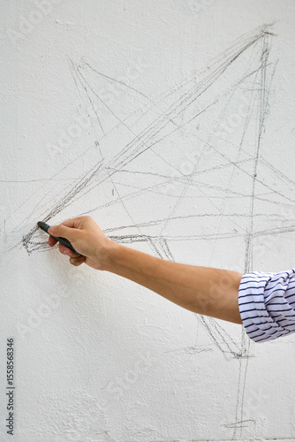 Young asian male with glasses sketches complex geometric shapes on white wall. Casual striped shirt, focused expression. Soft daylight illuminates scene