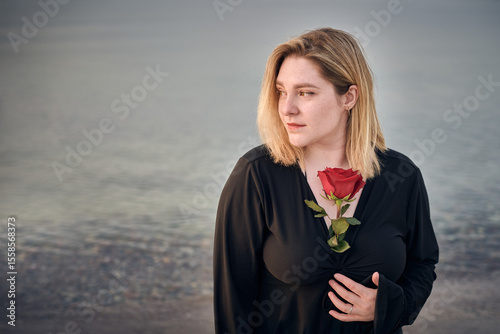 Young woman in black holds single red rose, gazing thoughtfully by serene waters. Mood evokes introspection and serenity, with backdrop of calming nature