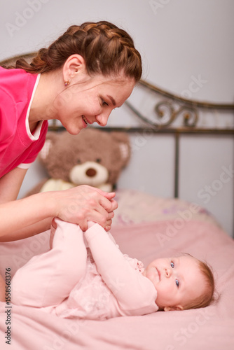 Young adult woman with braided brown hair joyfully engages with smiling baby girl on bed. Soft lighting enhances warm atmosphere. Background with teddy bear