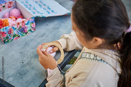 Young girl crafting with yarn in cozy home setting. Box of colorful yarns nearby. Bright lighting creates warm atmosphere. Focus on hands and materials