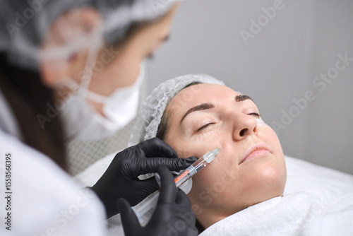 Young woman receives facial injection from professional in clinical environment. Procedure enhances aesthetics, showcasing precision, care, and serene atmosphere