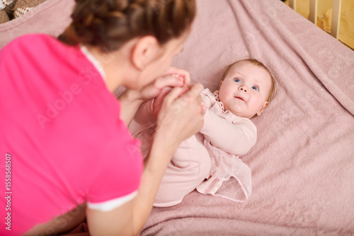 Smiling adult woman with brown hair interacts with baby girl. Baby lies on pink blanket. Warm lighting enhances soft pink colors, creating cozy atmosphere