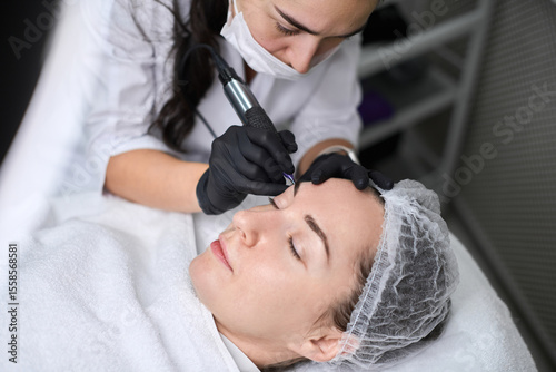 Young woman receives eyebrow permanent makeup in serene spa. Beauty technician, wearing gloves, focuses intently, creating calm, professional atmosphere