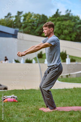 Young male performs open-arm yoga pose on grass in park. Wearing gray attire, focused and serene. Bright daylight, lush green backdrop, urban environment