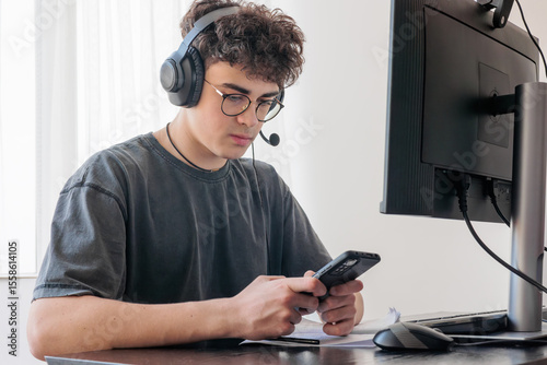 Young Male Student with Headphones Engaged in Online Learning Using Smartphone and Laptop — Teenager Studying at Home Preparing for Exams, Digital Education Concept. Copyspace