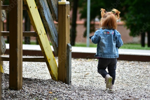 Red hair toddler girl playing in children playground in city park in summer. Little girl walking to climb stairs to slide down.