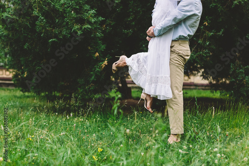 Caucasian groom playfully lifts bride in a white wedding dress on a green meadow, joyful outdoor wedding scene with authentic love and romantic atmosphere.
