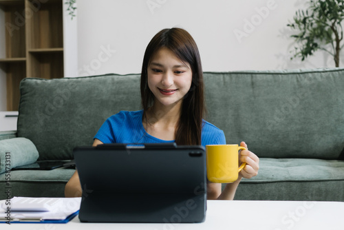 Smiling young Asian woman using digital tablet at home, enjoying remote work or online entertainment. Cozy lifestyle, modern tech habits, and casual home environment