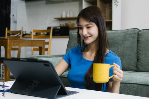 Smiling young Asian woman using digital tablet at home, enjoying remote work or online entertainment. Cozy lifestyle, modern tech habits, and casual home environment