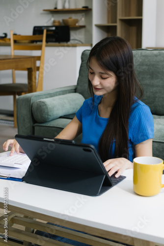 Smiling young Asian woman using digital tablet at home, enjoying remote work or online entertainment. Cozy lifestyle, modern tech habits, and casual home environment