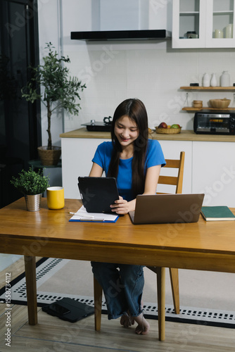 Smiling young Asian woman using digital tablet at home, enjoying remote work or online entertainment. Cozy lifestyle, modern tech habits, and casual home environment