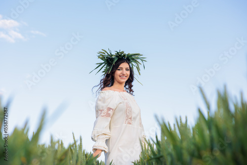 Beautiful Young Woman in Traditional Embroidered Dress and Green Wheat Wreath Looking at Camera in a Lush Rye Field.