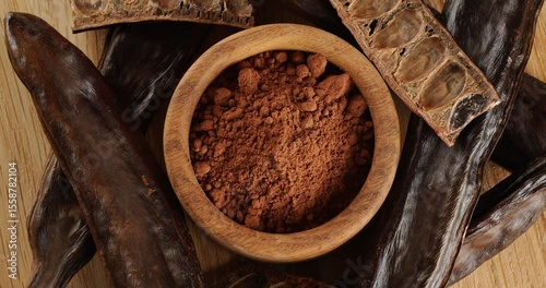 Carob pods and powder on a wooden board, top view, on a rotating table.