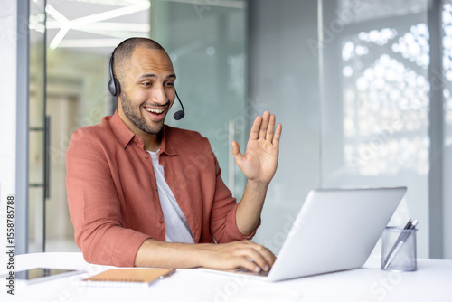 Support service worker with headset smiling, talking and consulting customers online remotely. Man working with laptop inside office.